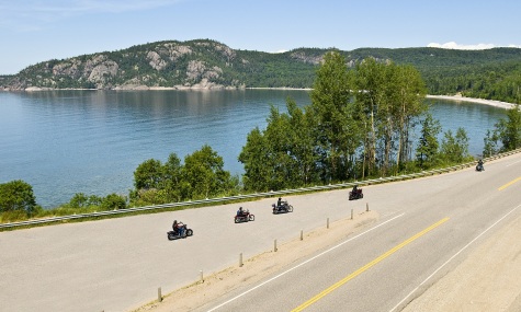 scenic view of  motorcycles riding down a winding road with water and cliff in the background