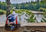 A motorcycle helmet with an american flag motif sitting on a cement wall with river and forest in the background