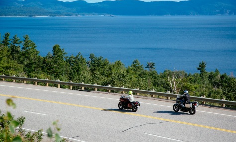 2 motorcycles riding down a scenic road with lake and hills in background