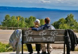 2 motorcycle riders sitting on a bench with grafitti overlooking scenic water view. motorcycle jackets are hanging on the end of the bench.